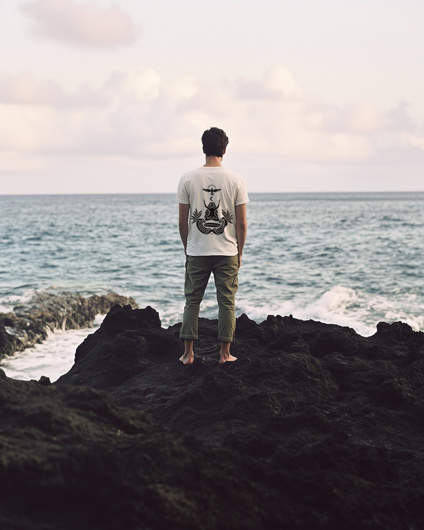 Back of the Goddess & Waves tee — man standing on lava rock facing the sea, the goddess mark printed in black