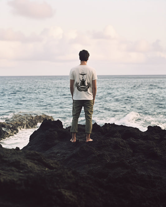 Back of the Goddess & Waves tee — man standing on lava rock facing the sea, the goddess mark printed in black